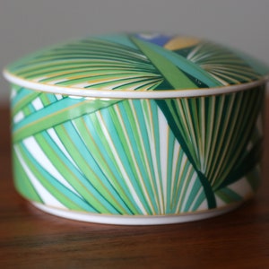 Puede incluir: Una caja redonda de porcelana blanca con un dise&ntilde;o de hojas de palma verdes, amarillas y azules. La tapa de la caja est&aacute; decorada con un patr&oacute;n similar.