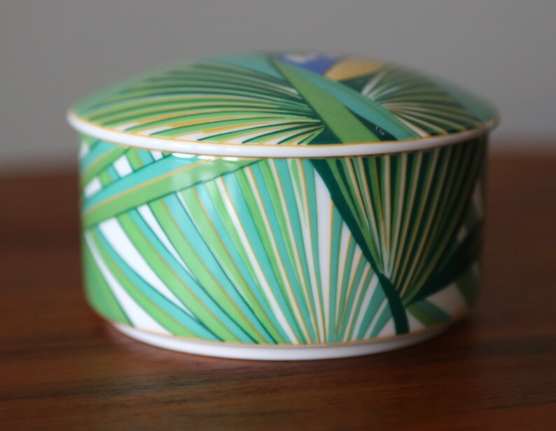Puede incluir: Una caja redonda de porcelana blanca con un dise&ntilde;o de hojas de palma verdes, amarillas y azules. La tapa de la caja est&aacute; decorada con un patr&oacute;n similar.