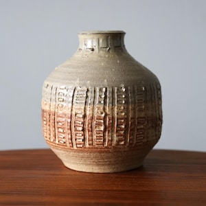 May include: A handmade ceramic vase with a textured finish. The vase has a rounded shape and a slender neck. The colour scheme includes beige, brown, and tan, with vertical textured patterns around the body.