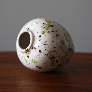 May include: White ceramic vase with a speckled brown and green glaze. The vase is round and has a wide opening.