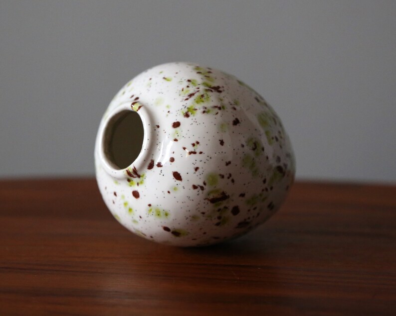 May include: White ceramic vase with a speckled brown and green glaze. The vase is round and has a wide opening.