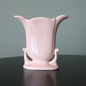 Peut inclure: Un vase en céramique rose clair avec un haut évasé et deux poignées incurvées. Le vase a une base de piédestal et des détails verticaux côtelés. La forme symétrique est élégante, adaptée aux compositions florales ou à l'exposition décorative.