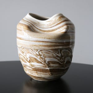 May include: A ceramic vase featuring a marbled pattern of white and earthy brown tones. The vessel has a rounded, organic shape with a wide, undulating rim that curves inward at the top, creating a distinct, asymmetrical silhouette with a smooth finish.