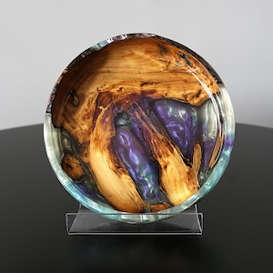 May include: A circular bowl crafted from natural wood and translucent purple resin. The wood features prominent grain patterns and organic edges, while the glossy resin fills the voids with a swirling, iridescent violet and teal color palette.