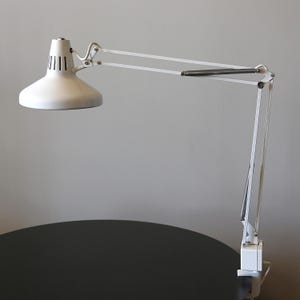 Könnte beinhalten: Weiße, verstellbare Schreibtischlampe mit konischem Schirm und Gelenkarm. Die Lampe hat einen Federmechanismus und einen rechteckigen Sockel. Die Lampe ist so konzipiert, dass sie verstellbar ist und eine gezielte Beleuchtung bietet.