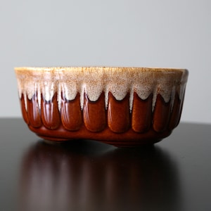 May include: A ceramic bowl with a fluted, scalloped exterior featuring a deep brown base colour that transitions into a lighter, textured beige glaze at the rim. The vessel has a rounded, elongated shape with a smooth, glossy finish throughout.