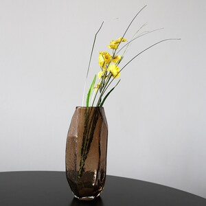 K&ouml;nnte beinhalten: Eine braune Glasvase mit strukturierter Oberfl&auml;che, die gelbe Blumen und lange gr&uuml;ne Stiele enth&auml;lt. Die Vase hat eine unregelm&auml;&szlig;ige, facettierte Form. Das Arrangement ist vor einem neutralen Hintergrund platziert und erzeugt eine schlichte, elegante Darstellung.