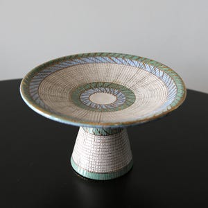 May include: A ceramic pedestal bowl featuring a wide, shallow basin and a tapered base. The surface is decorated with a grid pattern in white, light blue, and green, accented by fine, dark brown etched lines radiating from the center.