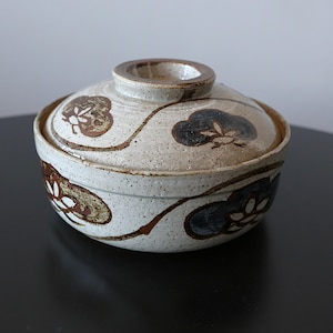 Könnte beinhalten: Eine Keramikschüssel mit Deckel und gesprenkelter, cremefarbener Oberfläche. Die Schüssel ist mit braunen und marineblauen Blumendesigns und einem braunen Linienmuster verziert. Der Deckel hat einen kleinen, kreisförmigen Griff.