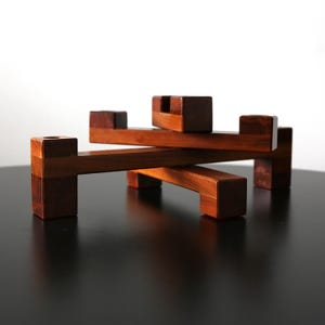 May include: A handcrafted wooden puzzle with a geometric design. The puzzle is made of dark brown wood and features interlocking pieces. The pieces are arranged in a layered, three-dimensional structure. The puzzle is displayed on a black surface.