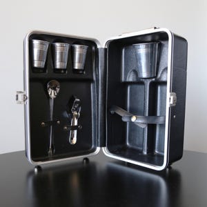 May include: Open black travel bar with silver metal shot glasses, a spoon, and a bottle opener. The interior is black with silver trim and a black leather strap. The bar is open, revealing the contents.