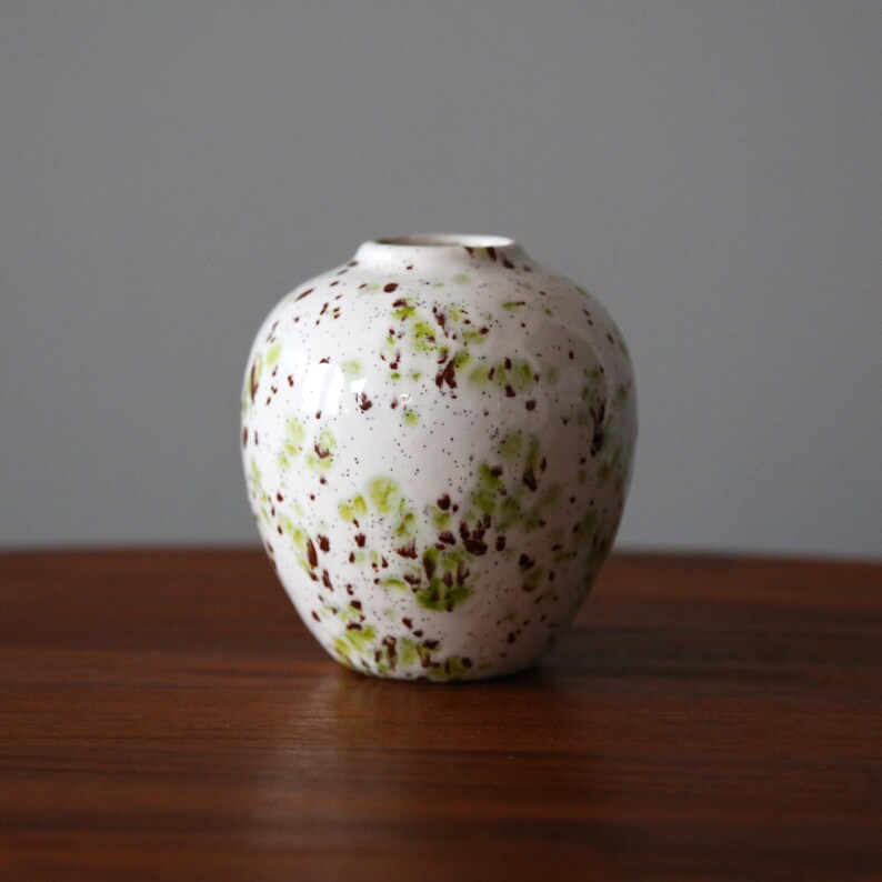 May include: A white ceramic vase with a speckled brown and green glaze. The vase has a rounded shape and a small opening at the top.