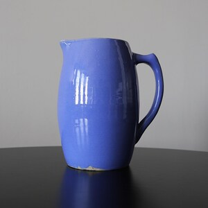 May include: A glossy periwinkle blue ceramic pitcher with a smooth, rounded body, a curved handle on the right side, and a small pouring spout at the top rim. The base of the pitcher shows a light gray, unglazed edge where the ceramic is exposed.