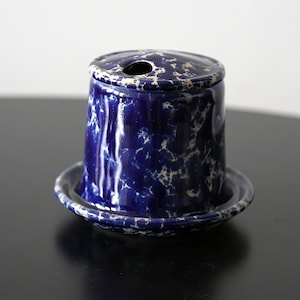 Pode incluir: Um porta-palitos de cerâmica azul e branca salpicada com tampa a condizer e uma pequena base de pires. O suporte cilíndrico tem um pequeno orifício no centro da tampa. O design lembra utensílios de cozinha vintage.