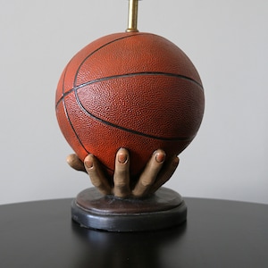 May include: A table lamp featuring a textured orange basketball resting in a sculpted hand base. The hand is painted in natural skin tones and sits atop a circular dark brown pedestal. A thin brass-coloured metal rod extends from the top.