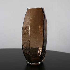 K&ouml;nnte beinhalten: Eine braune Glasvase mit unregelm&auml;&szlig;iger, strukturierter Oberfl&auml;che. Die Vase hat eine einzigartige, organische Form und ist f&uuml;r Blumen oder andere dekorative Gegenst&auml;nde konzipiert. Das Glas hat kleine Blasen, die f&uuml;r visuelles Interesse sorgen.