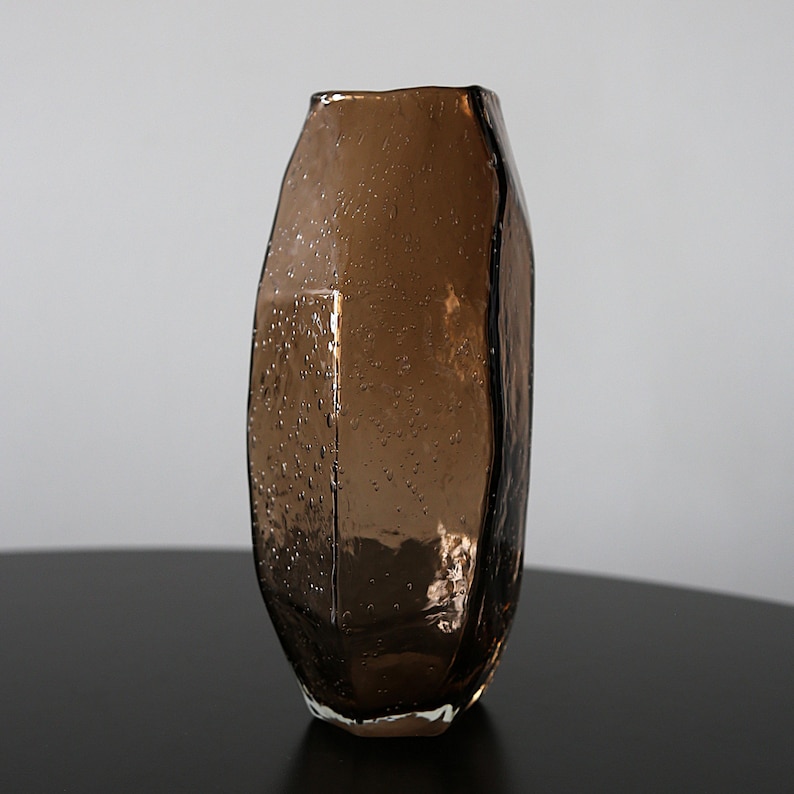 K&ouml;nnte beinhalten: Eine braune Glasvase mit unregelm&auml;&szlig;iger, strukturierter Oberfl&auml;che. Die Vase hat eine einzigartige, organische Form und ist f&uuml;r Blumen oder andere dekorative Gegenst&auml;nde konzipiert. Das Glas hat kleine Blasen, die f&uuml;r visuelles Interesse sorgen.