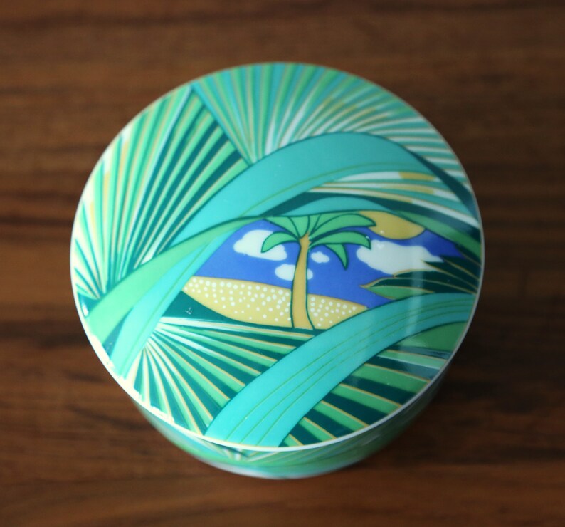 Puede incluir: Una caja de cer&aacute;mica redonda con un dise&ntilde;o verde, azul y amarillo de palmeras y una escena tropical.