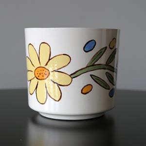 Peut inclure: Un cache-pot en céramique blanche orné d'un motif peint à la main représentant une grande marguerite jaune au cœur orange. Le dessin floral comprend une tige verte avec des feuilles et de petits accents ovales bleus et orange.