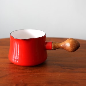 Puede incluir: Una pequeña cacerola de esmalte rojo con interior blanco y asa de madera. El asa está unida al lateral de la olla. La cacerola está sobre una superficie de madera. La olla está diseñada para cocinar y servir.