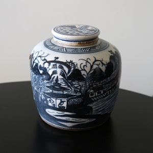 Puede incluir: Un jarrón de cerámica azul y blanco con tapa, que presenta una escena de paisaje pintada a mano. El jarrón tiene un diseño chino tradicional con un esquema de color azul y blanco.