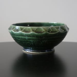 Pode incluir: Tigela de cerâmica verde com exterior canelado e borda ondulada. A tigela tem um acabamento brilhante e uma base azul escuro. A tigela é adequada para servir alimentos. O diâmetro da tigela é de aproximadamente 15 cm.