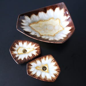 Puede incluir: Un conjunto de tres piezas de cerámica: una fuente rectangular y dos candelabros. Las piezas presentan un diseño marrón, blanco y amarillo. La fuente es rectangular con un borde ligeramente elevado. Los candelabros tienen un agujero central.