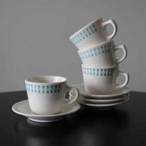 Puede incluir: Un juego de cuatro tazas de té de cerámica blanca con un patrón geométrico azul turquesa apiladas una encima de la otra. Las tazas están sobre una superficie negra con un platillo a juego en la parte delantera.