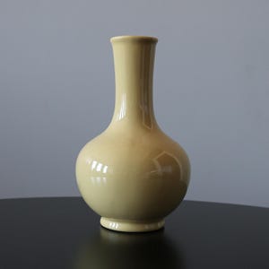 May include: A pale yellow ceramic vase with a rounded body and a long, slender neck. The vase has a glossy finish and sits on a small, circular base. The background is a neutral gray.