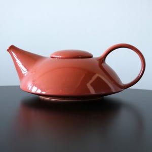 May include: A terracotta-colored ceramic teapot with a rounded body, a curved handle, and a small, circular lid. The teapot has a spout for pouring and sits on a dark surface. The design is simple and modern.