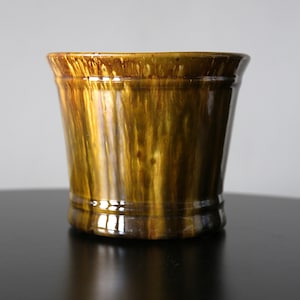 Op de afbeelding: Een cilindervormige keramische plantenpot met een glanzend, gevlekt bruin en goudgeel glazuur met verticale druippatronen. De pot heeft een licht uitlopende rand en een subtiele horizontale ribbel nabij de basis voor een gladde afwerking.