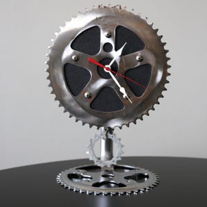 May include: A silver metal clock with a black face and red and white hands. The clock is made from repurposed bicycle parts and is mounted on a stand with two smaller gears.