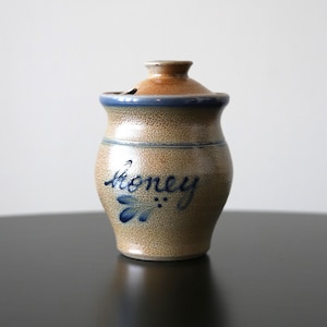 Puede incluir: Un tarro de miel de cerámica beige con tapa. El tarro tiene un borde azul y la palabra "honey" escrita en cursiva con un diseño floral azul. La tapa es marrón y tiene una pequeña abertura.