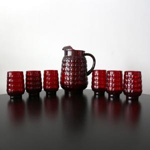 Può includere: Un set di sette bicchieri di vetro rosso e una caraffa abbinata. I bicchieri e la caraffa hanno un design testurizzato a forma di bolle.