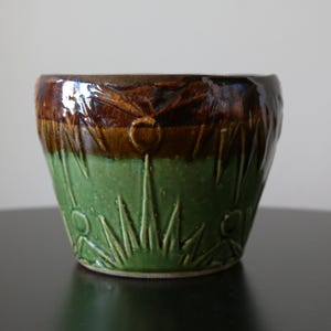 May include: A ceramic planter with a glossy finish. The top section is brown, the middle is a lighter brown, and the bottom is green. The planter has a textured design of flowers and blades of grass.