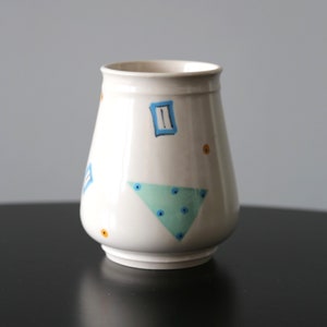 May include: White ceramic vase with a geometric design featuring blue, green, and yellow accents. The vase has a rounded base and a flared opening.