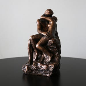 Op de afbeelding: Een bronzen beeld van twee figuren die elkaar omhelzen, zittend op een rotsachtige basis. Het beeld is een afbeelding van een romantisch stel.