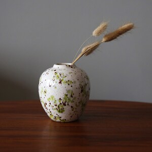May include: A white ceramic vase with green and brown speckles, holding dried beige grasses. The vase is on a dark brown wooden table.