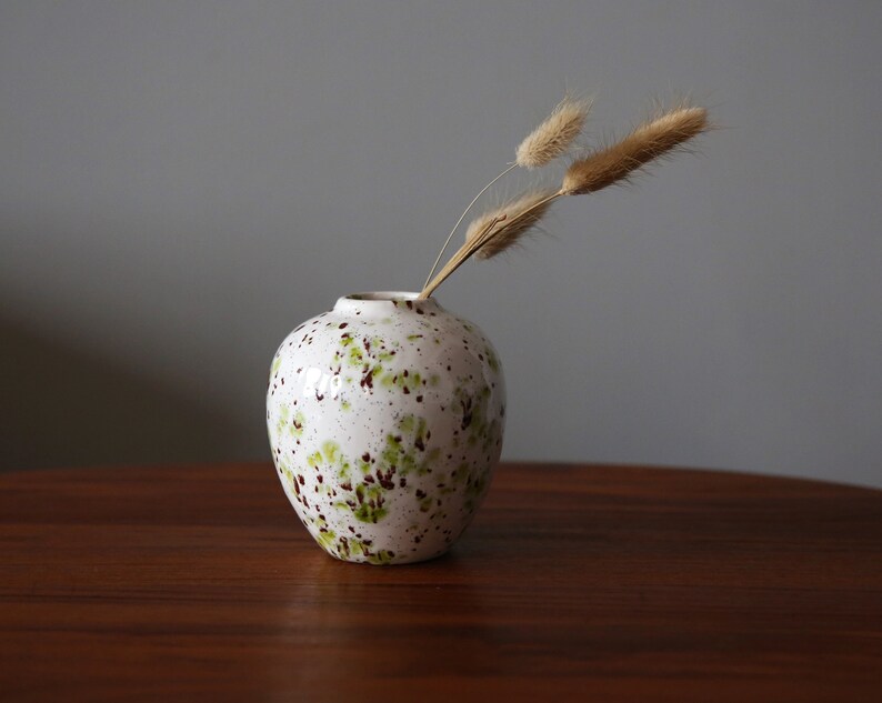 May include: A white ceramic vase with green and brown speckles, holding dried beige grasses. The vase is on a dark brown wooden table.