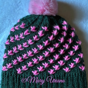 May include: A dark green knit hat with a pink floral pattern and a fluffy pink pom-pom on top. The hat has a ribbed brim and a textured design. The text "Mary Uriano" is visible on the lower part of the hat.