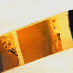 May include: A close-up of a strip of film with two frames showing a blurry image of a person in a yellow shirt. The film is slightly damaged and has a dark blue edge.