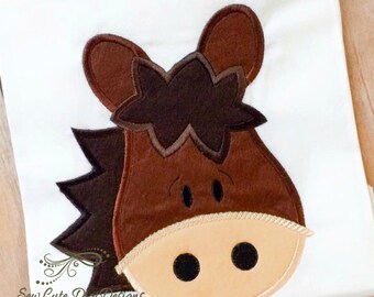 May include: A white t-shirt featuring a brown horse head applique with a dark brown mane and forelock, a light tan muzzle, and black embroidered eyes and nostrils. The text Sew Cute Digi Designs is printed in black in the bottom left corner.