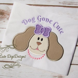 Può includere: Tessuto bianco con un cane ricamato marrone e bianco che indossa un fiocco viola e una collana di perline rosa. Il testo "Dog Gone Cute" è ricamato in viola sopra il cane.