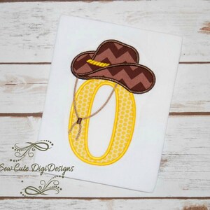 May include: Yellow embroidered number zero with a brown cowboy hat on top. The number has a polka dot pattern and the hat has a chevron pattern.