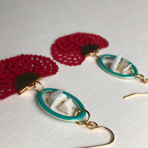 May include: A pair of red and gold earrings with a teal oval pendant featuring white shell pieces. The earrings are made with a delicate lace-like design.