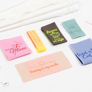 Puede incluir: Varias etiquetas de tela coloridas con textos como "Handmade by Mum" y "Wool is Cool". También incluye alfileres, una regla y tela blanca, todo sobre una superficie blanca.