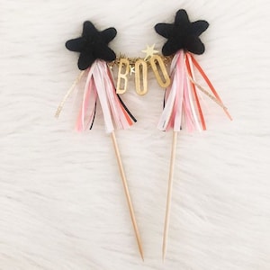 May include: Two black felt star cake toppers with gold glitter accents and a gold "BOO" banner. Each topper has pink, white, and red streamers attached to the bottom.