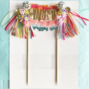 May include: A cake topper featuring the name "MARGOT" in gold letters, surrounded by pink, yellow and blue fringe. The topper also includes disco balls, smiley faces and flowers.