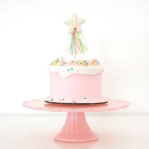 May include: A pink frosted cake with colorful sprinkles on a pink cake stand. The cake is topped with a white star-shaped topper with a rainbow streamer.