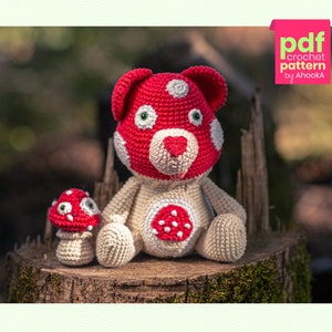 Può includere: Un orsetto fungo e un piccolo fungo all'uncinetto sono seduti su un ceppo d'albero. L'orso ha una testa a forma di cappello di fungo rosso con macchie bianche, un corpo beige e un fungo rosso sul petto. Il piccolo fungo è rosso con macchie bianche.